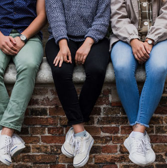 3 Jugendliche (weiblich/ männlich) auf einer Mauer dicht beieinandersitzend, die Beine sind zu sehen