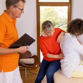 Bild während einer Funktionstherapie mit Arzt im orangen-farbigen Poloshirt und Patentin, die von Mitarbeiterin am Rücken behandelt wird