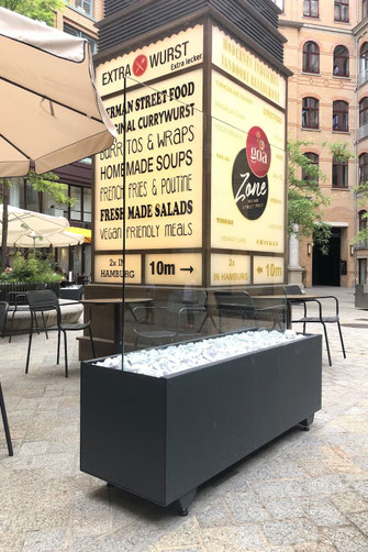 Casablanca Windschutz-Element mit dunkelgrauem Sockel, gefüllt mit weißen Dekosteinen und integriertem Glas-Windschutz in einem urbanen Umfeld.