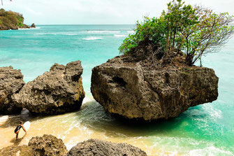 Woman standing on the beach with surfboard looking between rocks to the open turquoise sea.