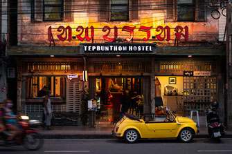 An old yellow car is parked in front of a run-down hostel in Bangkok.