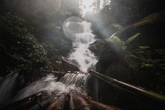 Langzeitbelichtung eines Wasserfalls mit einer kleinen Bambusbrücke im Vordergrund.
