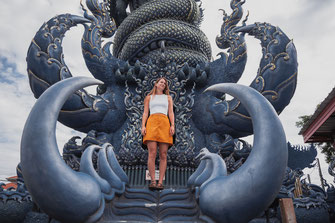 Eine Frau mit einen gelben Rock steht vor einer riesigen blauen Statue im Wat Rong Suea Ten.