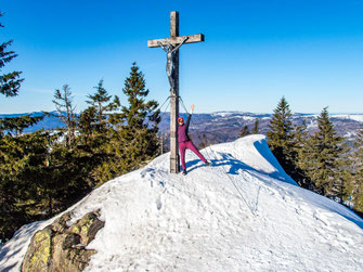 Wanderin am Gipfelkreuz des Großen Rachel bei einer Winterwanderung im Bayerischen Wald mit weiter Aussicht