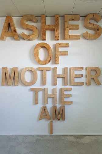 I AM THE MOTHER OF ASHES, von unten nach oben geschrieben, mit einer Gesamthöhe von etwa 3 Metern, hängen die Holzbuchstaben einzelnd an der Wand. Steht man davor, ist die Form eines Kriegsflugzeuges zu erkennen. Oliver Braig verunsichert oft und gerne mi