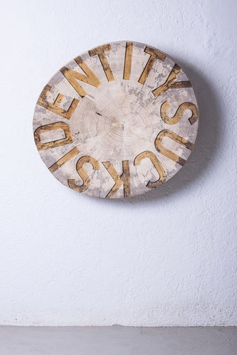 Die Holzskulptur Idenditysucks von Oliver Braig schwebt vor der Wand. Die Skulptur besteht aus einer Baumscheibe. Eine Scheibe aus Holz mit eingearbeiteten Großbuchstaben. Die Buchstaben reichen bis zum Rand der Scheibe. Die Skulptur ist lackiert.