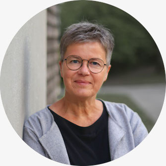 Porträt von Uschi Holstein, die entspannt an einer Wand lehnt, bis zur Brust sichtbar, vor unscharf grünem Hintergrund - einladend, freundlich und authentisch