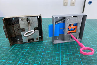 Auf einem Tisch mit grüner Schneidunterlage steht ein historisches Kastenschloss (Metall) mit Schlüssel. Rechts davon ein 3D-Druck des gleichen Kastenschlosses (Kunststoff). Dieses hat eine Öffnung um das Funktionsprinzip zu erkennen.