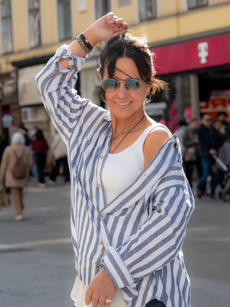 Lächelnde Frau mit Sonnenbrille auf der Mariahilferstrasse, sie trägt ein weisses Träger-Top unter einer blauweiss gestreiftem Leinenbluse.