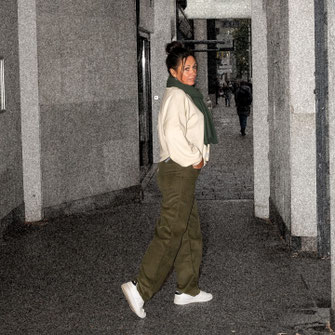 Renate in weissem Sweater, oliv-grünem Schal, oliv-grünen Hosen, weisen Sneakers in Hauseingang