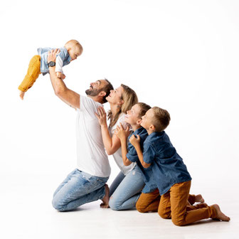 Photographie de famille professionnelle en studio dans l'Oise, montrant un père soulevant joyeusement son bébé en l'air, entouré de la mère et de leurs deux enfants alignés, style lumineux et complice par danimages.