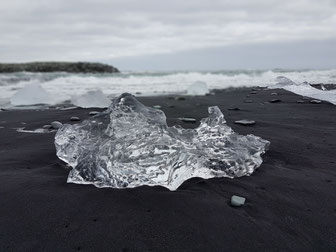 ein abgebrochenes Stück Gletschereis aus klarem Eis liegt auf schwarzem Lavasand. Dahinter leicht verschwommen, rollen die Meereswellen an den Strand