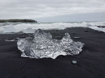 ein abgebrochenes Stück Gletschereis aus klarem Eis liegt auf schwarzem Lavasand. Dahinter leicht verschwommen, rollen die Meereswellen an den Strand ©My own Travel