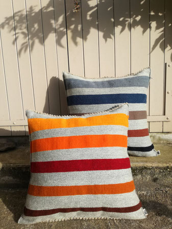 Deux grands coussins canapé à rayures exposés en plein air. Un coussin en rayures jaunes, oranges, marronnes, l'autre en rayures bleues, brunes et noires sur un fond de laine gris clair.