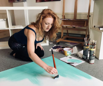 Portret van kunstenares Daphne Schrijver in haar atelier in Breda, omringd door kleurrijke en expressieve schilderijen. Daphne zit ontspannen op een kruk, gekleed in een witte blouse en lichtblauwe jeans, met opvallende rode hakken. 