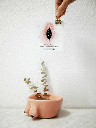 Das Bild zeigt einen getöpferten Blumentopf mit Brüsten, darin ist eine kleine Eukalyptuspflanze. Über dem Topf hält eine Hand eine Postkarte mit einer Zeichnung einer Vulva und dem Spruch "May the Power of all Yonis be with you"