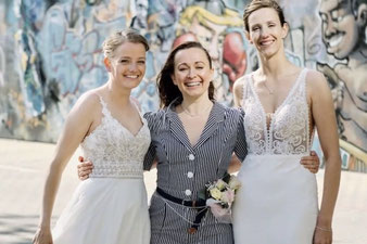 Constanze Marienfeld steht strahlend lachend zwischen ihren beiden Bräuten die beide weiße Brautkleider tragen und ebenfalls strahlen. 