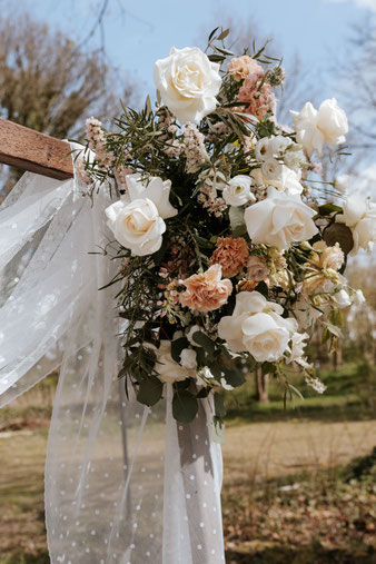 oppulente Blumendeko mit weißen Rosen, aprikotfarbenen Nelken und Eukalyptus am Traubogen. Der weiß gepunktete Stoff am Traubogen weht im Wind