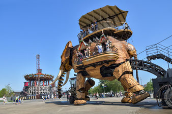 Elefant aus Stahl und Holz auf "Les Machines de l'Île" in Nantes mit Passagieren, im Hintergrund ein Karussell