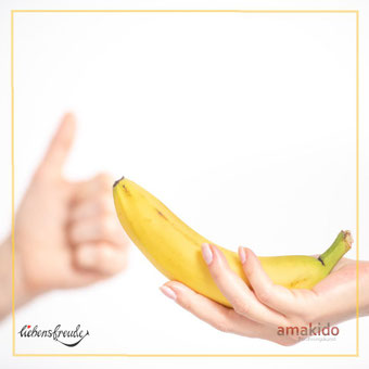 Hand hält eine Banane; im Hintergrund eine Daumen-hoch-Geste – Metapher für achtsame Berührung; feiner Rahmen; Logos: Liebensfreude links unten, amakido rechts unten.