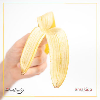 Aufgeschälte Banane wird in der Hand präsentiert – Metapher, dass für achtsame Berührung etwas fehlt; feiner Rahmen; Logos: Liebensfreude links unten, amakido rechts unten.