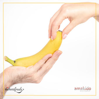 Banane in einer Hand; andere Hand berührt sanft die Spitze – Metapher für achtsame Berührung; feiner Rahmen; Logos: Liebensfreude links unten, amakido rechts unten.