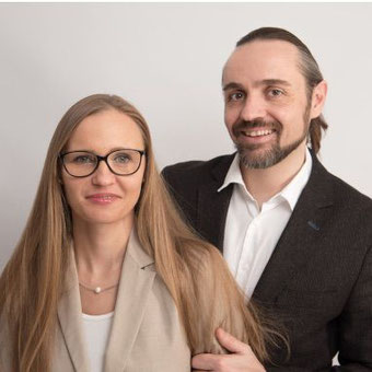 zwei Personen nebeneinander vor hellem Hintergrund; links Person mit langen, glatten Haaren, Brille, beigem Blazer und Perlenkette, rechts Person mit zurückgebundenen Haaren, Bart, weißem Hemd und dunklem Sakko, lächelnd – Team Liebensfreude