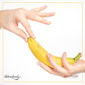 Zwei Hände führen eine präzise Finger-Geste an der Bananenspitze aus – Metapher für achtsame Berührung; feiner Rahmen; Logos: Liebensfreude links unten, amakido rechts unten.