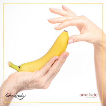 Banane ruht in einer Hand; zweite Hand nähert sich der Spitze mit feiner Geste – Metapher für achtsame Berührung; feiner Rahmen; Logos: Liebensfreude links unten, amakido rechts unten.