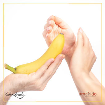 Banane in der Hand; daneben eine schützende-Geste – Metapher für achtsame Berührung und Grenzen/Konsens; feiner Rahmen; Logos: Liebensfreude links unten, amakido rechts unten.