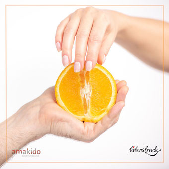 Hände halten eine halbierte Orange; eine Hand berührt die Schnittfläche – Metapher für achtsame Berührung; mit Logos: amakido links unten, Liebensfreude rechts unten.