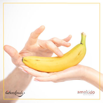 Banane auf geöffneter Handfläche; darüber eine schwebende, sanfte Finger-Geste – Metapher für achtsame Berührung; feiner Rahmen; Logos: Liebensfreude links unten, amakido rechts unten.