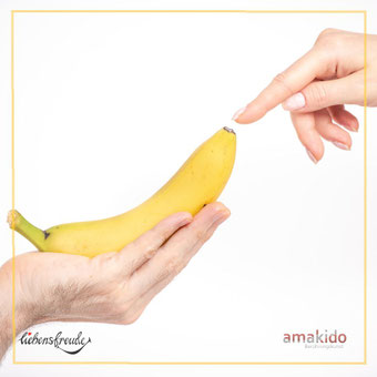 Hand hält eine Banane; ein Finger nähert sich der Spitze – Metapher für achtsame Berührung; feiner Rahmen, heller Hintergrund; Logos: Liebensfreude links unten, amakido rechts unten.