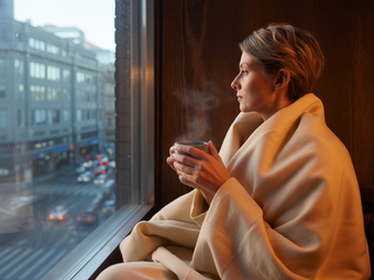Frau sitzt am Fenster, erschöpft und eingekuschelt in eine Decke. Sie hält eine Tasse Tee in der Hand und blickt auf den Großstadtverkehr runter. 