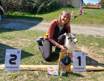 Christine kniet neben ihrem Hund Finn, der vor einem Schild mit der Zahl '1' sitzt, umgeben von Hundeleckerlis und einem Pokal im Freien.