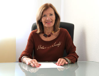 Margit Königsecker, Coach & Trainerin - sitzt an einem Glasschreibtisch, trägt braunes Oberteil, Hände am Tisch vor einem weißen Blatt Papier