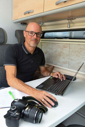 Ein Mann sitzt in einem Campervan am Tisch. Vor ihm auf dem Tisch steht ein Laptop-Computer. Auf dem Tisch liegen eine Kamera und ein Notizbuch.