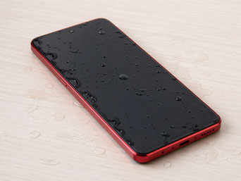 淡い木目のテーブルの上に置かれた赤いAndroidスマートフォンの画面に、水滴が広がって付着している様子を写した写真。水濡れや水没による不具合をイメージしやすいクローズアップ画像。