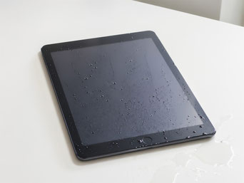 白いテーブルの上に、画面と本体に水滴が付着した黒いiPadが置かれている写真。端末の周囲にも水が広がっており、iPadの水濡れや水没トラブルを表した状態イメージ。