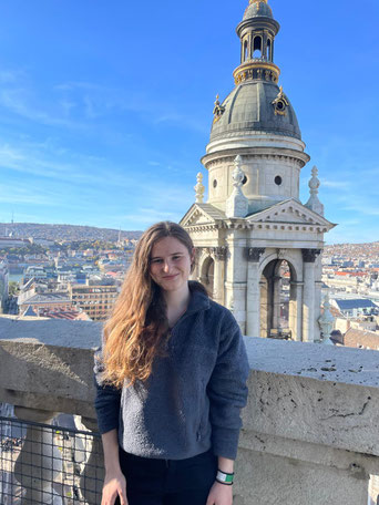 Johanna steht vor einer Mauer auf einem hohen Gebäude, hinter ihr ist der Turm eines historischen Gebäudes zu sehen und die Umrisse einer Ortschaft.