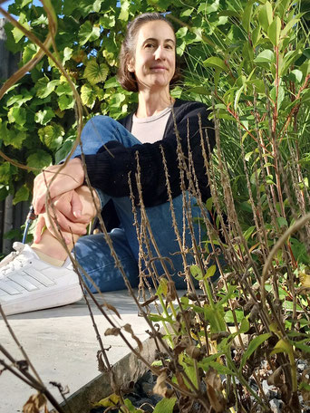 Michaela Dauscher sitzt auf einer Mauer im Garten, ein Bein angezogen, die Hände liegen auf der Wade. Hinter ihr wächst eine Weinpflanze an einer Wand hoch, vor ihr sind auch grüne Pflanzen. Sie lächelt.