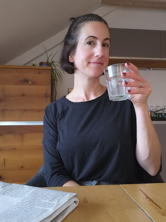 Michaela Dauscher sitzt am Tisch, trinkt aus ihrem Glas Zitronentee, vor ihr liegt eine Zeitung, im Hintergrund ist ihre Holzküche zu erkennen