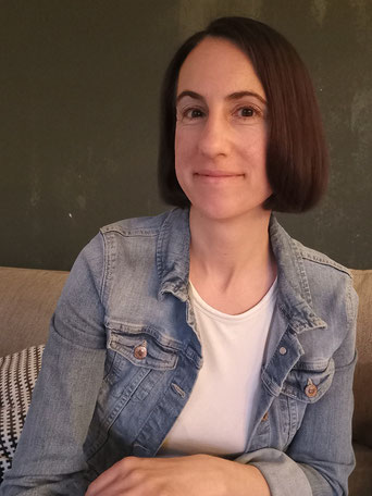 Michaela Dauscher sitzt auf dem Sofa, hellblaue Jeansjacke, weißes T-Shirt drunter. Die Wand dahinter ist dunkelgrün.