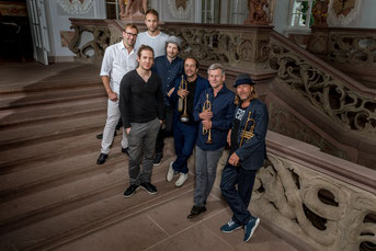 7 Männer stehen auf einer barocken Treppe. Drei halten Trompeten in der Hand.