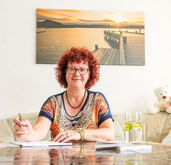 Ines Illing sitzt am Tisch, an der Wand ein Bild mit einem Steg am See