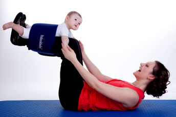 Mutter liegt auf dem Rücken. und streckt die Beine in die Luft. Ihr Baby ist mit einem Gurt an den Beinen der Mutter gesichert. Dabei Trainiert die Mutter ihren Körper. Beide haben Spaß.
