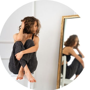 Eine junge Frau sitzt nachdenklich auf einem Hocker und blickt in einen Spiegel, der ihr Bild reflektiert. Die Szene wirkt ruhig und introspektiv.