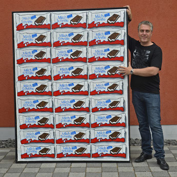 Künstler Divo Santino steht neben einem großformatigen XXL-Gemälde, das eine serielle Anordnung von Kinder Milchschnitte Packungen in kräftigen Acrylfarben zeigt.