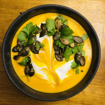 Orangene Kürbissuppe in grauer Schale auf Holz. Auf der Suppe sind geröstete braune Kürbiskerne und frische grüne Petersilie zu sehen. Außerdem ist weißer Kürbisjoghurt in schönen Mustern in die Orangene Suppe eingearbeitet.