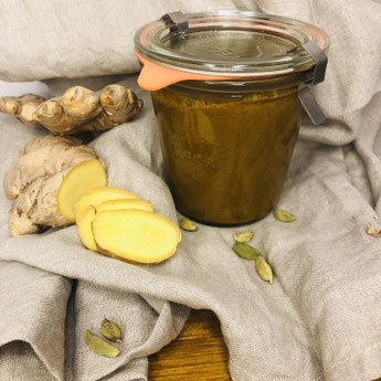 Goldene Milch Paste im Weck-Glas. Sie ist golden, gelblich, braun. Dekoriert in Beiges Leinentuch, auf dem aufgeschnittener Ingwer und grüne Kardamomkapseln liegen.